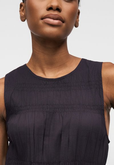 Femme portant un haut noir sans manches avec un détail froncé au niveau de la poitrine et un col rond.