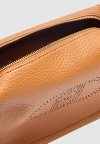 Tote tas van bruin leer met een gestructureerd oppervlak, gouden rits en een reliëf logo detail aan de binnenzijde. Compact ontwerp met een gladde voering.