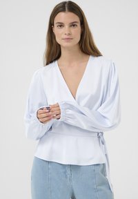 Svetlomodrá blúzka s previazaním vyrobená z hladkej látky. Má hlboký výstrih do V, balónové rukávy s elastickými manžetami a detail viazania v páse.