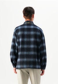 Uomo che indossa una camicia a maniche lunghe a scacchi blu scuro e nera e pantaloni beige chiaro, girato di spalle su uno sfondo bianco semplice.