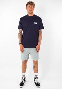 Camiseta de manga corta azul marino con logo turquesa en el pecho, acompañada de pantalones cortos gris claro, zapatillas de caña alta negras y calcetines estampados.