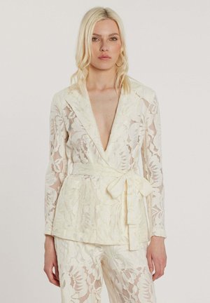 Donna bionda che indossa un blazer in pizzo color crema con cintura annodata in vita e pantaloni in pizzo abbinati, in piedi davanti a uno sfondo neutro.