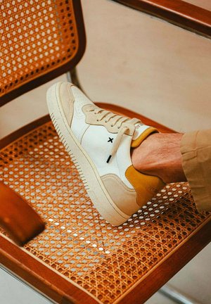 Weiße und beige Sneaker mit einer strukturierten Oberseite, glattem Zehenbereich und Gummisohle. Mit gelbem Akzent an der Ferse und schwarzem Logo-Detail.