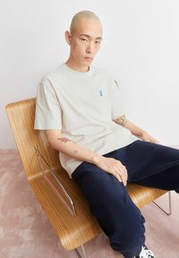 Lichtbeige T-shirt met een blauw lintlogo, gecombineerd met donkere marineblauwe broek. Het model zit op een gebogen houten stoel met een strak ontwerp.