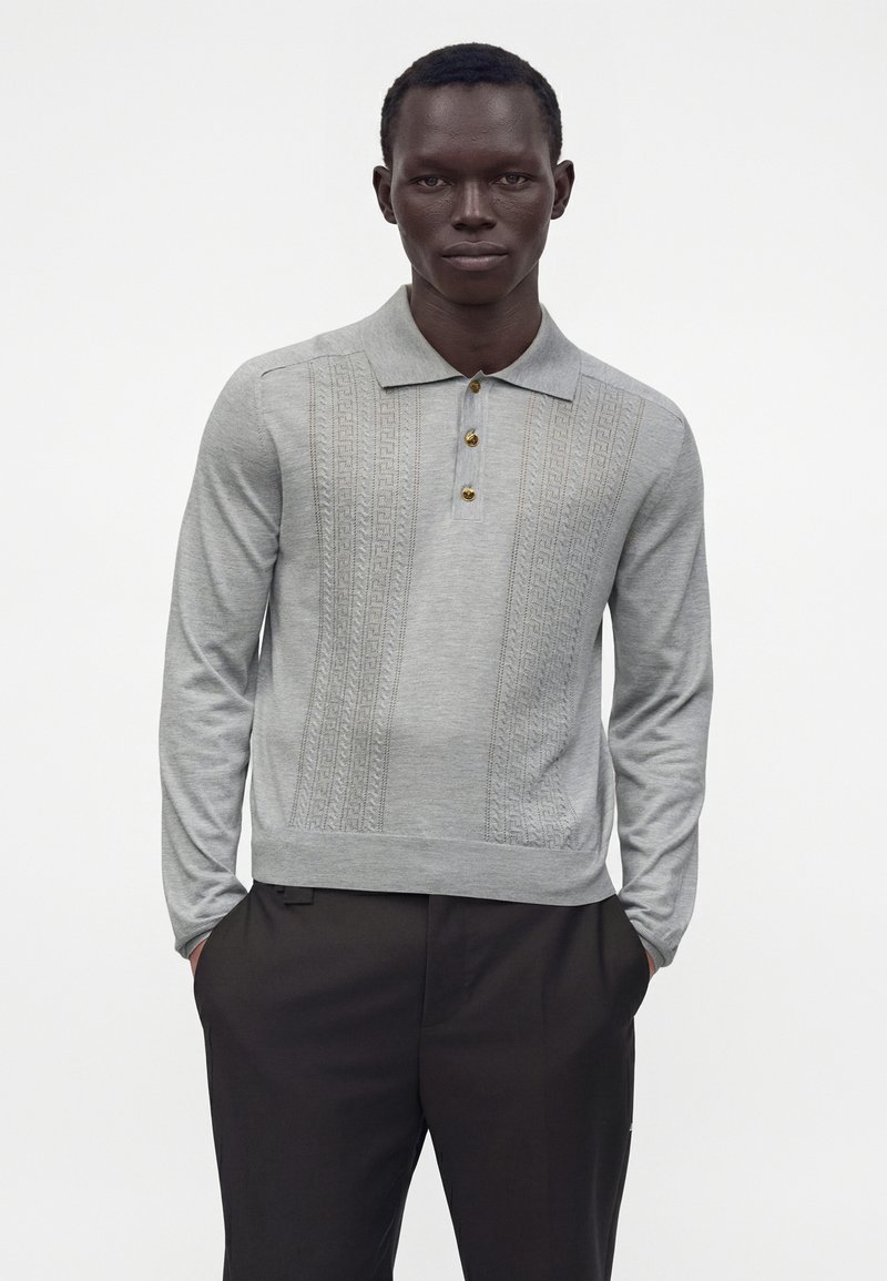 Homme portant un polo en maille gris clair à manches longues avec un devant à motif et des boutons dorés, associé à un pantalon noir, mains dans les poches.