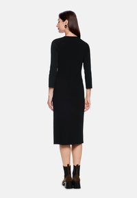 Schwarzes, figurbetontes Kleid mit Dreiviertelärmeln, knielang, getragen mit braunen Stiefeln mit Absatz und runden schwarzen Ohrringen, von hinten gezeigt.