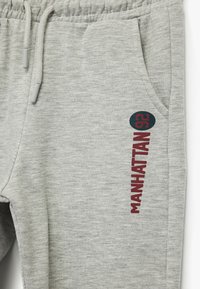 Grå joggerbukser i et blødt materiale, med en sidelomme og rød "MANHATTAN" tekst langs benet med en rund teal-farvet accent.