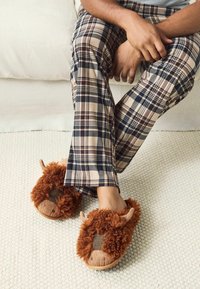Brune, fuzzy tøfler med øredetaljer, plaiderede stof accenter, båret med rulluvede bukser på et tekstureret beige tæppe.