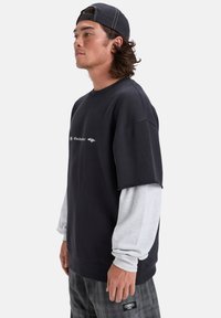 Felpa nera oversize con maniche lunghe grigie sotto, caratterizzata da un logo sul davanti. Il tessuto appare morbido; vestibilità casual e rilassata.