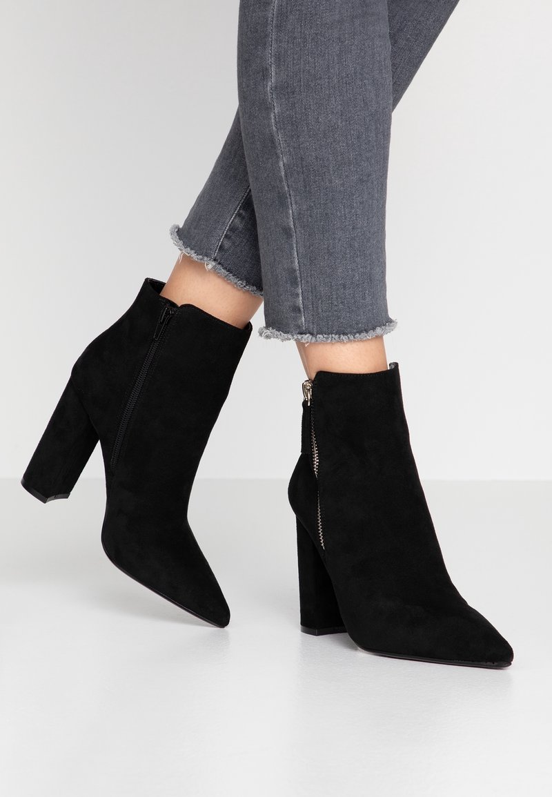Ankle boots neri in suede con punta appuntita, tacco alto a blocco e dettaglio con cerniera sul lato. Indossati con jeans grigi con orlo consumato.