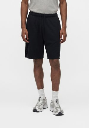 Mężczyzna ubrany w czarne, sięgające do kolan szorty ze sznurkiem, szarą koszulkę, białe skarpety do kostek oraz srebrne buty sportowe Nike, stojący na białym tle.