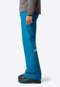 Pantaloni da esterno turchesi con vestibilità rilassata, realizzati in materiale impermeabile; presentano il logo del marchio sulla gamba sinistra e sono abbinati a stivali neri.