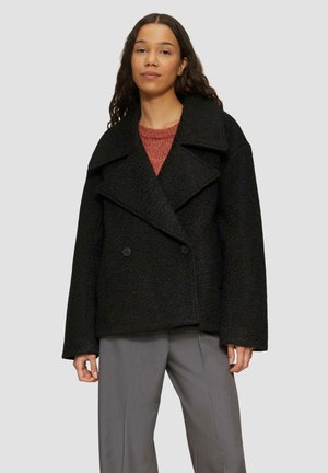 Manteau noir croisé avec grands revers, tissu texturé et deux boutons à l'avant, associé à un pantalon gris et un pull rouge.