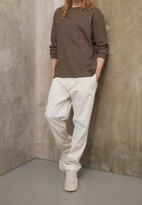 Hnědá žebrovaná mikina s dlouhými rukávy, kombinovaná s volnými bílými kalhotami a světlými teniskami. Jednoduchý a moderní design.