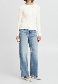 Biały kardigan w prążki z okrągłym dekoltem i zapięciem na guziki, w połączeniu z jasnoniebieskimi spodniami o szerokich nogawkach i białymi butami z ostrym noskiem.