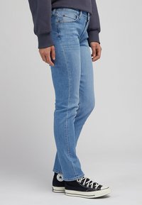 Vaaleansiniset farkut, joissa on kapea malli, viisi taskua, kultainen viimeistely ja keskimääräinen vyötärö, yhdistettynä mustiin tennareihin.