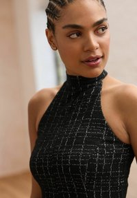 Femme aux cheveux tressés portant un haut noir sans manches à col haut avec un motif à carreaux subtil, regardant légèrement vers sa droite.