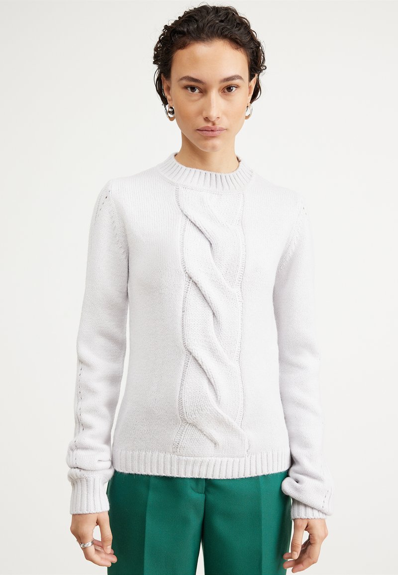 Maglione bianco lavorato a maglia con un design a trecce texture, colletto e polsini a coste, e dettagli di cucitura laterale abbinato a pantaloni verdi.