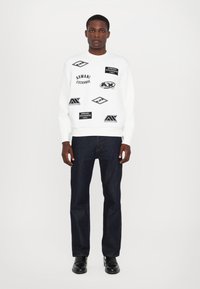 Biała bluza z czarnymi graficznymi logo i tekstem, w połączeniu z ciemnymi dżinsami. Obuwie to czarne, błyszczące, sznurowane buty.