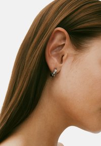 Svart hoopörhänge med en vriden design, visat på en modells öra. Örhänget har en slät, glänsande yta och minimal synlig hårdvara.