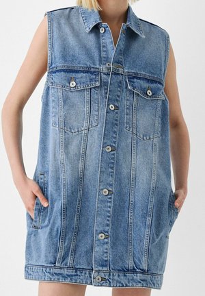 Veste en jean bleu clair sans manches avec boutons sur le devant, deux poches poitrine à rabat et poches latérales, portée par une personne les mains dans les poches.