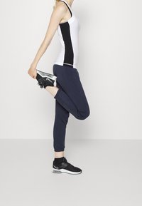 Vit och svart tanktopp, marinblå svettbyxor och svarta sportskor. Personen sträcker ut ett ben bakåt, håller om ankeln.