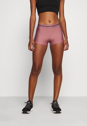 Kobieta ubrana w liliowe spodenki kompresyjne Under Armour, czarny sportowy biustonosz oraz czarne buty sportowe, stojąca z rękami swobodnie opuszczonymi wzdłuż ciała.