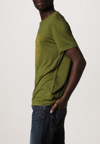 Olivengrøn kortærmet t-shirt med rund halsudskæring og sideslidsdetaljer, prydet med et gult grafisk design på forsiden.