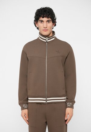 Homme se tenant debout, portant une veste zippée marron avec des rayures blanches au col et à l'ourlet, assortie à un pantalon marron, devant un fond uni.