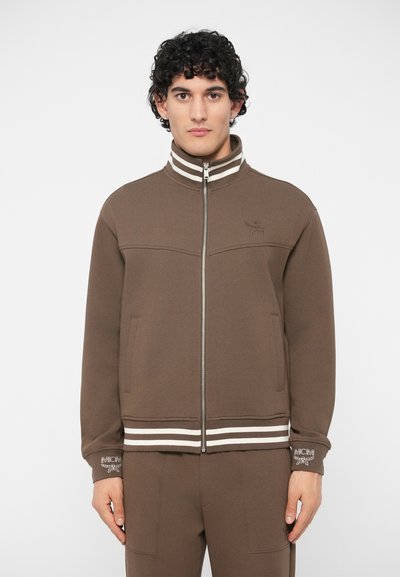 Chaqueta marrón con cremallera hecha de tejido suave, con un cuello alto, rayas blancas en el cuello y el dobladillo, y bordado del logo de MCM.