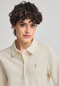 Krämfärgad långärmad polotröja i mjukt tyg, med krage och fem knappar. Broderad logotyp på bröstet.
