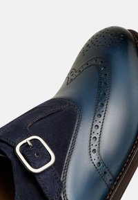Marineblaue Lederschuhe mit einem Wildlederakzent, präsentiert mit einem Broguemuster, glatten und strukturierten Oberflächen sowie einem silbernen Schnallendetail.