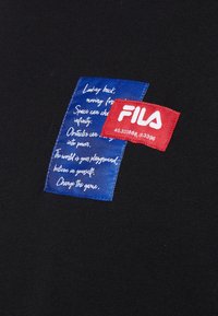 Tessuto nero con due etichette: un logo FILA rosso e un'etichetta blu con testo bianco che presenta frasi ispiratrici.