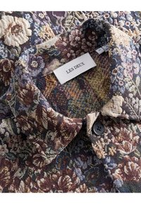 Camisa con estampado floral que presenta una tela texturizada en tonos burdeos, azul marino y crema, con un botón negro y una etiqueta de marca.