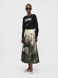 Γυναίκα φοράει μαύρο μακρυμάνικο τοπ Moschino, γυαλιά ηλίου και μπεζ-μαύρη φούστα με σχέδιο πρόβατα, στέκεται σε απλό φόντο.