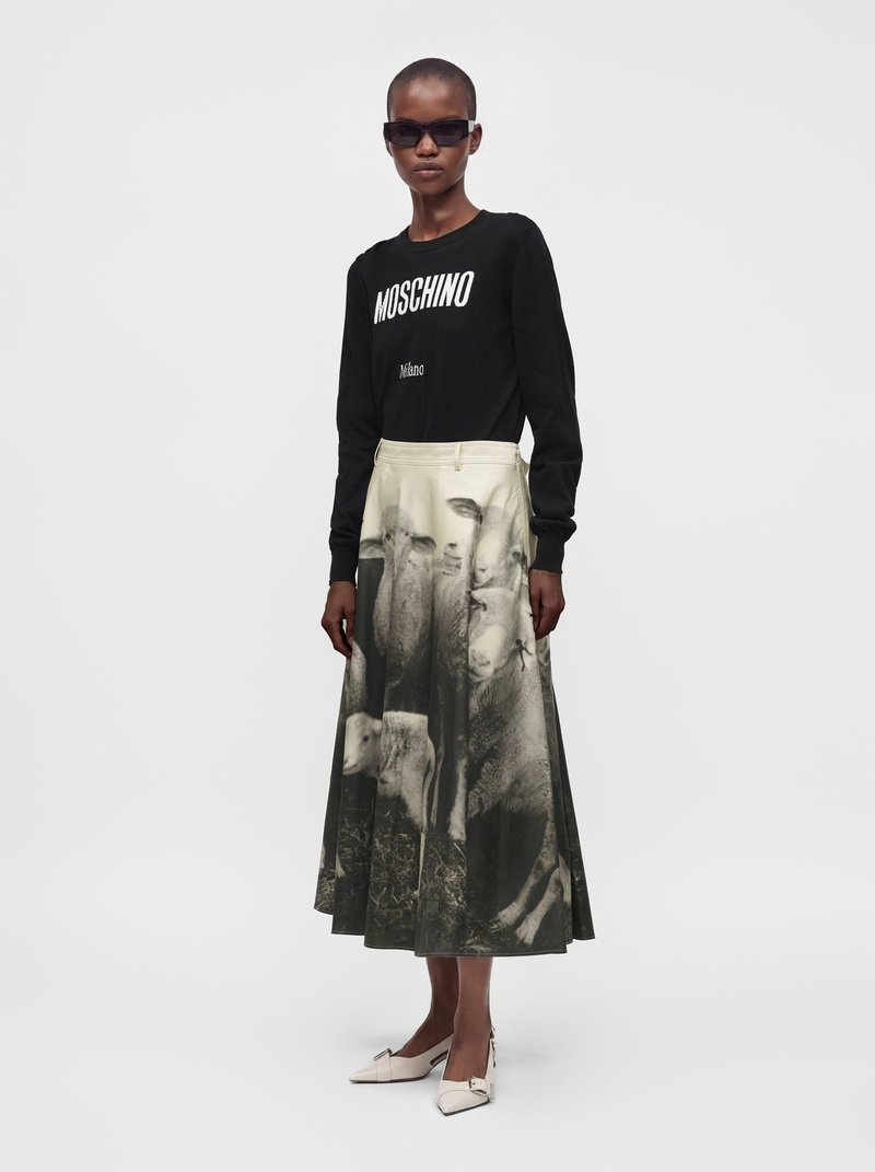 Femme portant un haut Moschino noir à manches longues, des lunettes de soleil et une jupe beige et noire avec imprimé moutons, debout devant un fond uni.