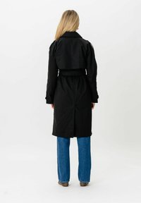 Femme blonde portant un trench-coat noir ceinturé et un jean bleu, debout de dos à la caméra sur un fond blanc.