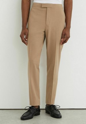 Man draagt beige op maat gemaakte broek en zwarte leren veterschoenen, staand tegen een witte muur.