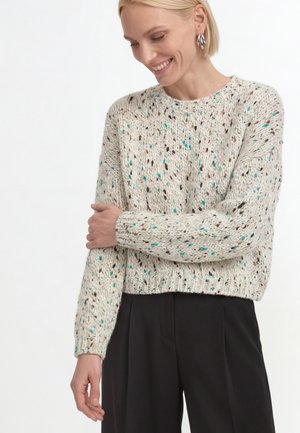 Lächelnde Frau mit kurzen blonden Haaren, die einen cremefarben gesprenkelten Strickpullover und schwarze Hosen trägt, hält ihren linken Arm mit der rechten Hand.