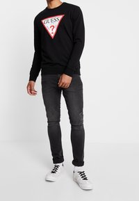 Man in een zwarte Guess-sweater met rood logo, donker gewassen jeans, witte sokken en witte sneakers, staand tegen een simpele witte achtergrond.