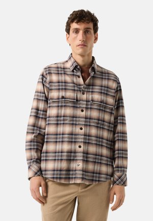 Ung mand iført en beige, navy og brun ternet skjorte med knapper over en brun undertrøje og beige kostbukser, stående mod en ensfarvet baggrund.