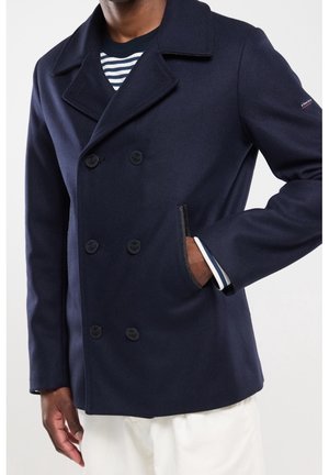 Homme portant un caban bleu marine à double boutonnage avec les mains dans les poches, sur une chemise rayée et un pantalon blanc, sur un fond uni.