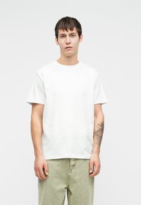 Λευκή βαμβακερή μπλούζα με κοντά μανίκια και στρογγυλή λαιμόκοψη. Το μοντέλο φοράει επίσης ανοιχτό πράσινο τζιν παντελόνι, αποκαλύπτοντας ένα τατουάζ στον πήχη.