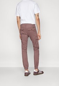 Heren cargobroek in een stoffig rozekleur; voorzien van elastische manchetten, achterzakken met kleppen en een taps toelopende pasvorm. gedragen met witte sneakers.