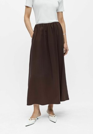 Frau trägt ein weißes kurzärmeliges Oberteil und einen langen braunen Rock mit Taschen, kombiniert mit weißen spitzen Slingback-Schuhen.