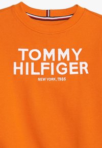 Oranžový svetr s kulatým výstřihem a bílým vyšitým textem "TOMMY HILFIGER" a menším "NEW YORK, 1985" pod názvem značky.