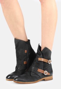 Botas negras hasta el tobillo con un upper texturizado, correas de cuero marrón con acentos metálicos, punta afilada y un tacón corto de madera apilada.