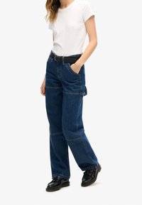 Jeans de mezclilla anchos de color azul oscuro con costuras en contraste, bolsillos laterales y un cinturón negro. Combinados con una camiseta blanca de manga corta.