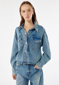 Chaqueta de mezclilla azul clara con cierre de botones, dos bolsillos en el pecho y mangas largas. Presenta un cuello clásico y un corte relajado.