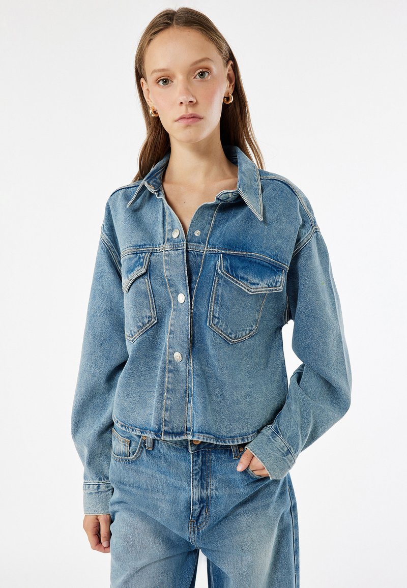 Chaqueta de mezclilla azul clara con cierre de botones, dos bolsillos en el pecho y mangas largas. Presenta un cuello clásico y un corte relajado.